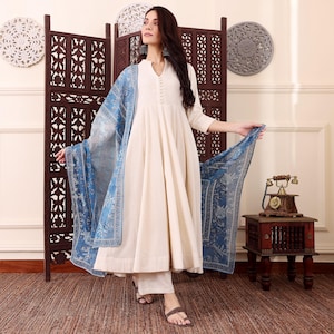 May include: A cream-colored, long-sleeved dress with a flowing skirt and matching pants. A blue patterned scarf is draped over the shoulders. The outfit is paired with brown sandals. The background includes a decorative screen and wall decor.