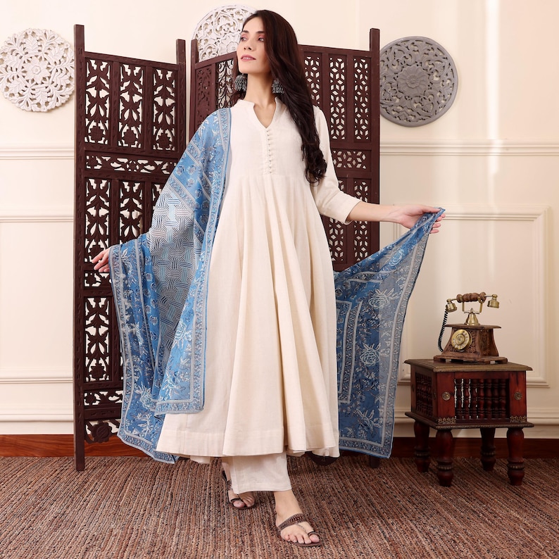 May include: A cream-colored, long-sleeved dress with a flowing skirt and matching pants. A blue patterned scarf is draped over the shoulders. The outfit is paired with brown sandals. The background includes a decorative screen and wall decor.