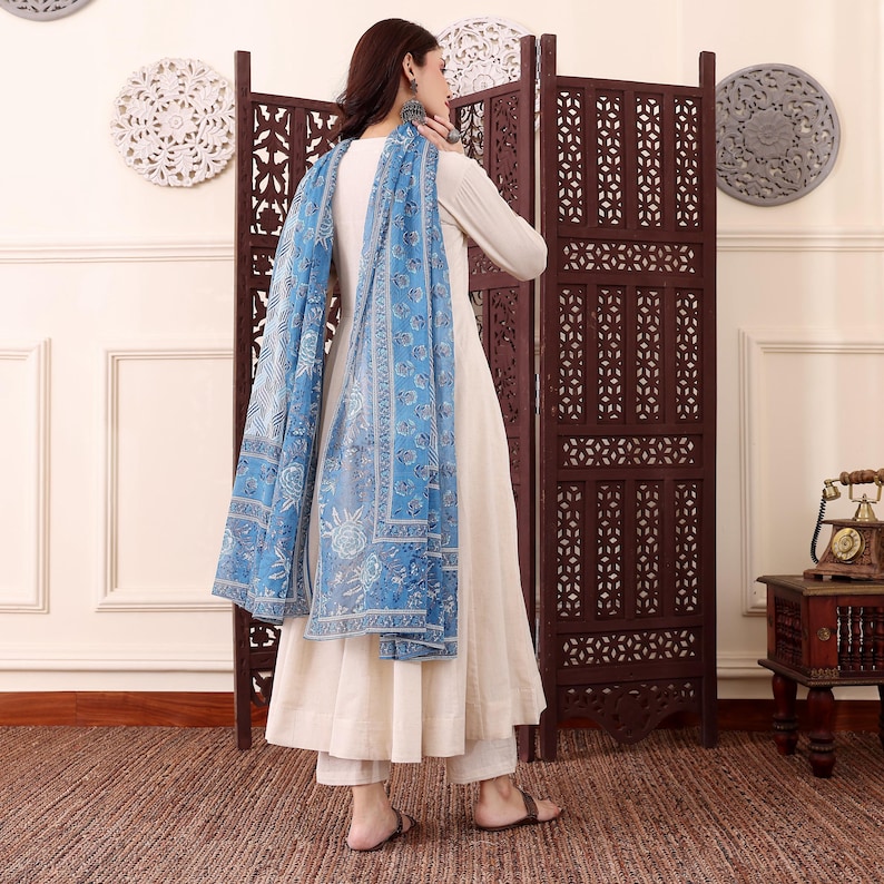 May include: An off-white long-sleeve dress and matching pants are paired with a blue patterned scarf. The scarf features a floral design and drapes over the shoulders. The outfit is worn with sandals.