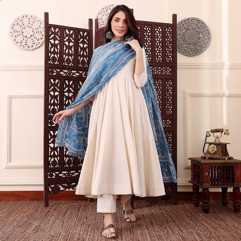 May include: An off-white, flowing dress with a matching pair of pants and a blue patterned shawl. The outfit is worn with brown sandals. A wooden room divider and decorative wall hangings are in the background.