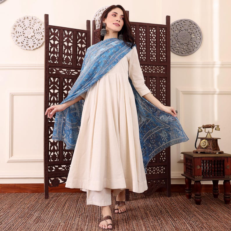 May include: A cream-colored, long-sleeved dress with a flowing skirt and matching pants. A blue patterned scarf is draped over the shoulders. The outfit is worn with sandals. The background includes a wooden screen and decorative wall hangings.