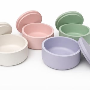Puede incluir: Conjunto de seis pequeños recipientes redondos con tapas en colores pastel: blanco, rosa, verde y lila. Las tapas están ligeramente elevadas y los recipientes están dispuestos sobre una superficie blanca.