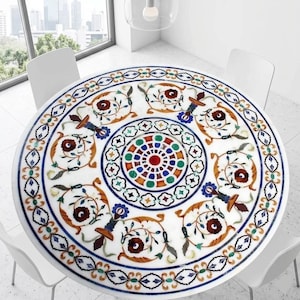 May include: A round white marble table with an intricate inlaid floral design in shades of brown, blue, green, and red. The table is surrounded by white chairs and set in a bright, modern room. The table's diameter is approximately 48 inches.