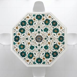 May include: An octagonal white marble table with a floral inlay design in teal, gold, and blue. The table is surrounded by three white chairs. The table's intricate pattern is centered with a starburst design.