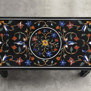 May include: A rectangular black marble coffee table with a floral inlay design. The table features a white border and a central circular design with colorful flowers in red, blue, yellow, and white. The table is supported by four black legs.