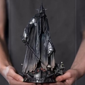 May include: A detailed black statue of a figure in armor, holding a sword. The figure wears a helmet with a tall, pointed crown and a long, tattered cloak. The statue is on a detailed base with spikes and other decorative elements.