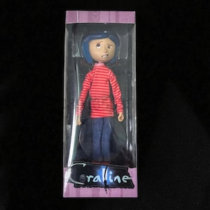 May include: A doll of a character from the movie "Coraline" is displayed in a clear plastic box. The doll has blue hair, a red and white striped shirt, and blue jeans. The box has the word "Coraline" printed on it.