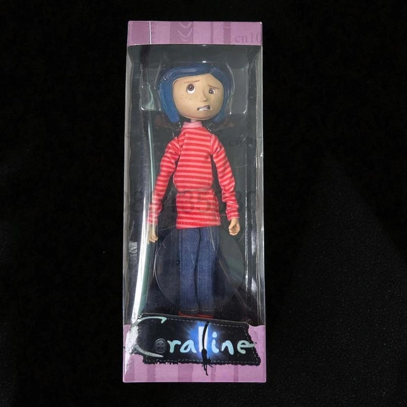 May include: A doll of a character from the movie "Coraline" is displayed in a clear plastic box. The doll has blue hair, a red and white striped shirt, and blue jeans. The box has the word "Coraline" printed on it.
