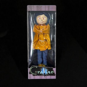 May include: A doll of a character from the movie "Coraline" is displayed in a clear plastic box. The doll has blue hair, freckles, and is wearing a yellow raincoat, blue jeans, and yellow boots. The box has the word "Coraline" printed on it.