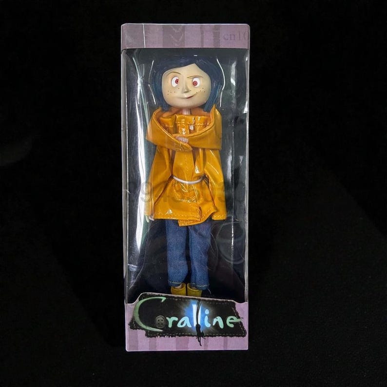 May include: A doll of a character from the movie "Coraline" is displayed in a clear plastic box. The doll has blue hair, freckles, and is wearing a yellow raincoat, blue jeans, and yellow boots. The box has the word "Coraline" printed on it.