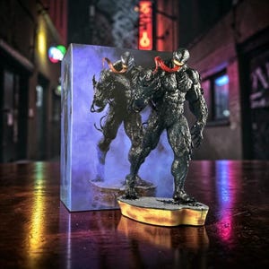 May include: A black Venom figurine with a red tongue, standing on a yellow and gray base. The figurine is in front of a box with a mirrored image of the figure. The background is a blurred city scene.