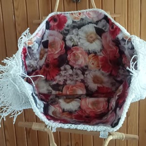 K&ouml;nnte beinhalten: Eine wei&szlig;e geh&auml;kelte Tragetasche mit floralem Innenfutter. Die Tasche hat einen gewebten Griff und einen dekorativen Fransenbesatz an den Kanten. Der Innenstoff zeigt ein Muster aus roten, rosa und wei&szlig;en Blumen.