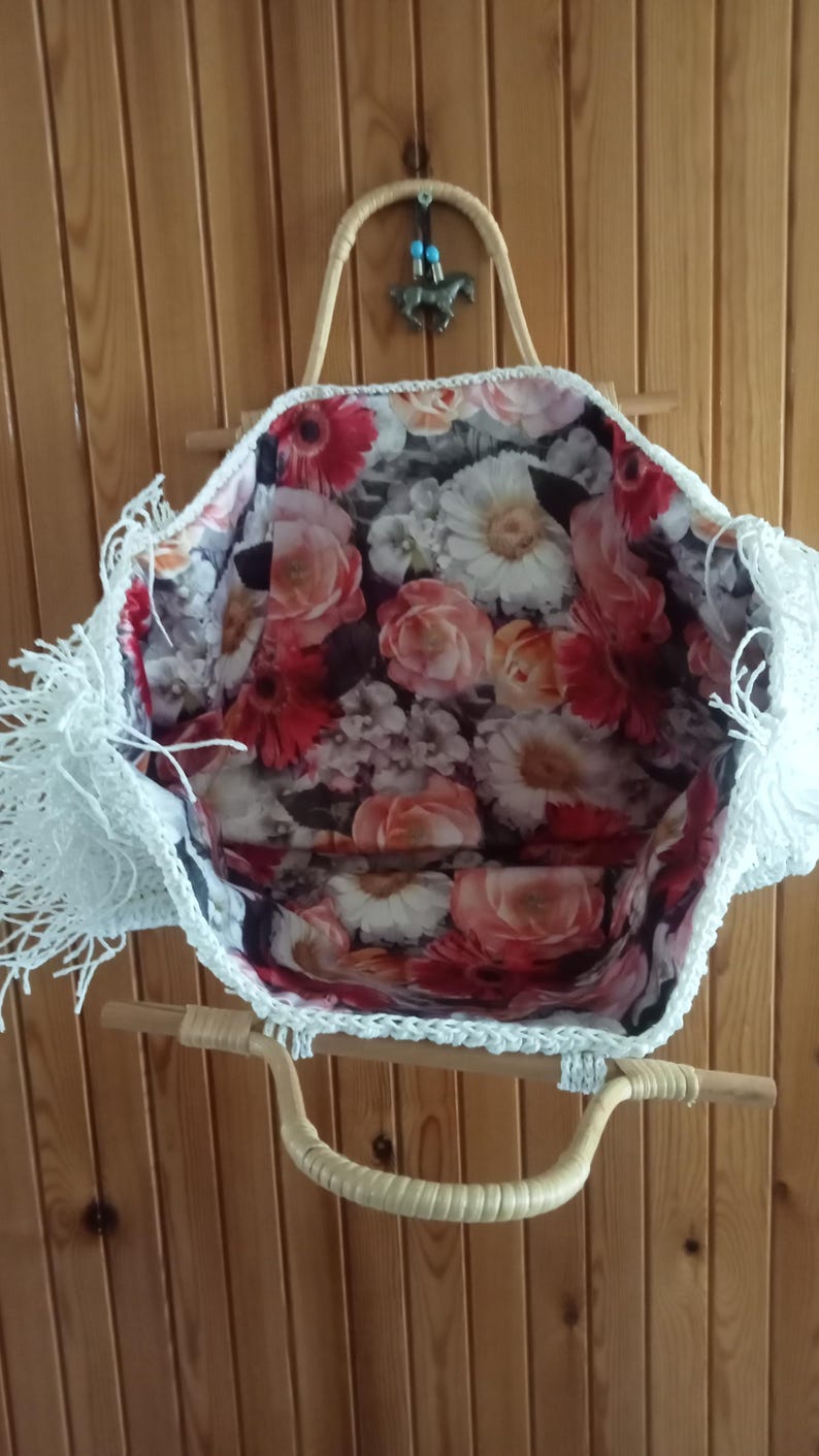 K&ouml;nnte beinhalten: Eine wei&szlig;e geh&auml;kelte Tragetasche mit floralem Innenfutter. Die Tasche hat einen gewebten Griff und einen dekorativen Fransenbesatz an den Kanten. Der Innenstoff zeigt ein Muster aus roten, rosa und wei&szlig;en Blumen.