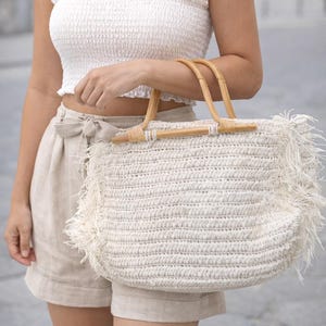 K&ouml;nnte beinhalten: Eine cremefarbene, gewebte Tragetasche mit Bambusgriffen und Fransen. Die Tasche wird von einer Person getragen, die ein wei&szlig;es Crop-Top und beige Shorts tr&auml;gt. Eine goldene Halskette ist ebenfalls zu sehen.