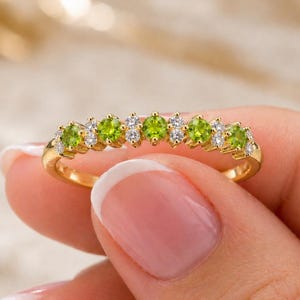 May include: A gold ring featuring a row of round, light green gemstones alternating with small, clear gemstones. The ring is held by a person with a light skin tone.