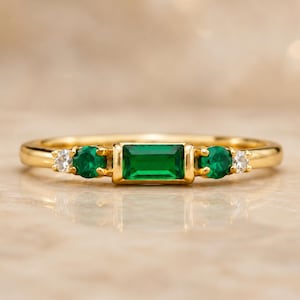 Peut inclure: Une bague en or ornée d'une émeraude rectangulaire au centre, flanquée de deux émeraudes rondes et de deux petits diamants. La bague a un anneau délicat et une finition polie, mettant en valeur le vert vif des émeraudes.