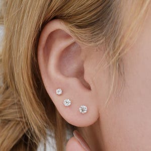 Peut inclure: Gros plan sur une oreille ornée de trois boucles d'oreilles en diamant de tailles différentes. La plus petite est en haut, la taille augmentant vers le bas. Les boucles d'oreilles scintillent. L'oreille est sur un fond flou.