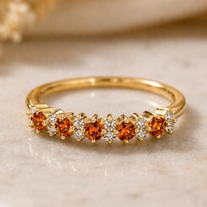 May include: A gold ring featuring a row of orange gemstones alternating with small white gemstones. The ring has a thin band and is set against a neutral background. The gemstones are round and evenly spaced.
