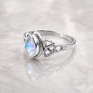 Peut inclure: Une bague en argent avec une pierre précieuse irisée de coupe marquise. La bague présente un motif de nœud celtique sur le bracelet et autour du sertissage de la pierre précieuse. La bague est présentée sur une surface réfléchissante.