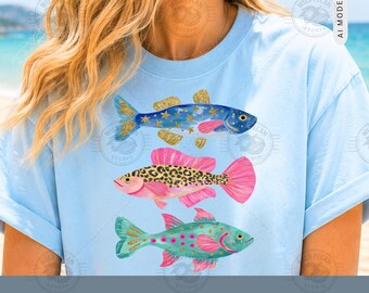 Pode incluir: Uma t-shirt azul-clara com uma estampa gráfica de três peixes empilhados verticalmente. O peixe de cima é azul com estrelas douradas, o do meio tem um padrão de leopardo rosa e preto, e o peixe de baixo é verde-azulado com pontos rosa e estrelas.
