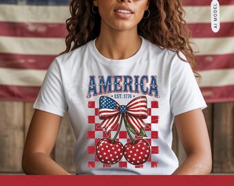 Pode incluir: Uma t-shirt branca de manga curta com um gráfico de duas cerejas com padrões de estrelas, rematadas com um laço patriótico. O texto AMERICA e EST. 1776 aparece acima das cerejas, sobre um design de fundo aos quadrados vermelhos e brancos.