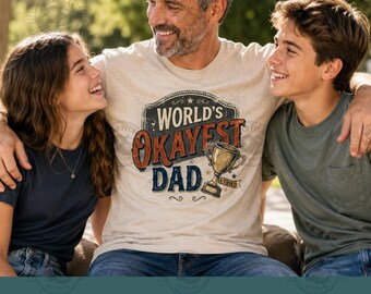 Pode incluir: Uma t-shirt bege de manga curta com uma impressão gráfica com o texto "WORLD'S OKAYEST DAD" em letras laranja e azuis, acompanhada por uma ilustração de um troféu dourado e o texto "I TRIED" numa faixa por baixo do troféu.