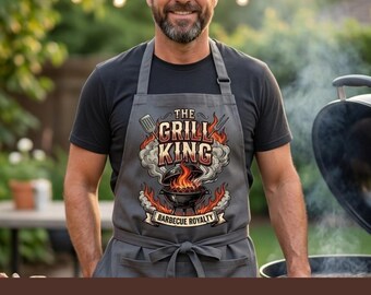 Pode incluir: Um avental cinzento com um gráfico de um grelhador a carvão com chamas e fumo, com o texto THE GRILL KING e BARBECUE ROYALTY impresso numa fonte ousada e estilizada, acompanhado por ilustrações de uma espátula e um garfo de churrasco.
