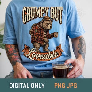 Könnte beinhalten: Ein hellblaues T-Shirt mit einer Grafik eines braunen Bären, der eine Mütze und ein rot kariertes Hemd trägt und eine Tasse mit der Aufschrift Grumpy Juice hält. Der Text lautet Grumpy But Loveable. Das Design zeigt ein braunes Banner und einen Rucksack.