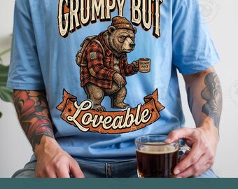 Pode incluir: Uma t-shirt azul-clara com o gráfico de um urso castanho a usar um gorro e uma camisa de xadrez vermelha, segurando uma caneca com a etiqueta Grumpy Juice. O texto diz Grumpy But Loveable. O design inclui uma faixa castanha e uma mochila.