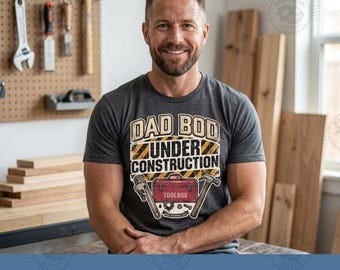 Pode incluir: Uma t-shirt de manga curta cinzento-escura com o texto DAD BOD UNDER CONSTRUCTION em letras brancas e amarelas, com um gráfico de uma caixa de ferramentas vermelha, uma chave de canos e um martelo na parte frontal da peça.