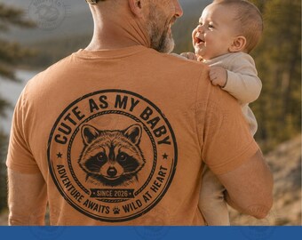 Pode incluir: Uma t-shirt laranja com um gráfico circular preto nas costas. O design inclui uma ilustração de um guaxinim, o texto "CUTE AS MY BABY", "SINCE 2026", "ADVENTURE AWAITS" e "WILD AT HEART", juntamente com um pequeno ícone de pata.