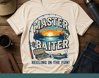 Pode incluir: Uma t-shirt de cor creme com um gráfico de uma cana de pesca, uma caixa de pesca, um pequeno barco e um lago ao pôr do sol. O texto diz MASTER BAITER e REELING IN THE FUN! O design inclui um peixe a saltar e pinheiros no fundo.