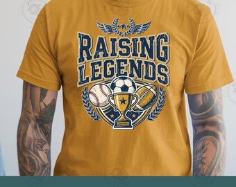 Pode incluir: Uma t-shirt amarelo-mostarda com uma estampa gráfica com o texto RAISING LEGENDS em azul escuro. O design inclui uma bola de basebol, uma bola de futebol, uma bola de futebol americano e um troféu dentro de uma moldura de louros.