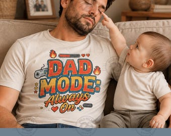 Pode incluir: Uma camiseta bege com o texto "DAD MODE: Always On" em vermelho, laranja e azul. A camisa apresenta um gráfico de controle de videogame e um design de barra de carregamento. Um bebê está estendendo a mão para a pessoa que está usando a camisa.