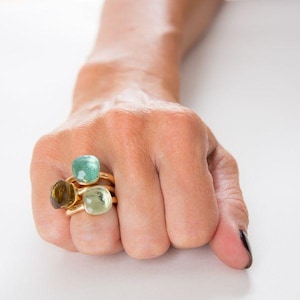 Può includere: Tre anelli in oro con pietre preziose lisce e arrotondate in stile cabochon nei toni del verde acqua traslucido, verde chiaro e marrone scuro. Gli anelli sono impilati, ognuno con una fascia in metallo lucido e una singola pietra arrotondata.