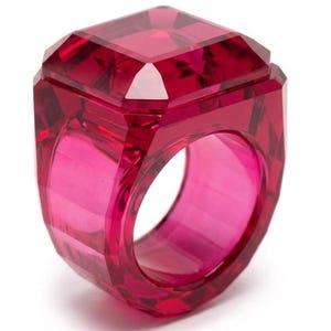 Puede incluir: Un anillo grande, facetado, de color rojo rubí. El anillo tiene una parte superior cuadrada y una banda gruesa. El material transparente permite el paso de la luz, creando un accesorio vibrante y llamativo. El diseño es geométrico y audaz.