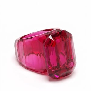 Puede incluir: Un gran anillo facetado de color rojo rubí con una piedra central de talla esmeralda. El anillo está hecho de un material transparente, y las facetas capturan la luz, creando un efecto brillante. El anillo está sobre un fondo blanco.