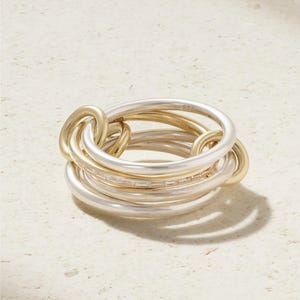 Könnte beinhalten: Ein mehrreihiger Ring aus ineinandergreifenden silber- und goldfarbenen Reifen. Einer der silbernen Reifen ist mit einer Reihe kleiner, klarer Baguette-geschliffener Steine besetzt, die das Licht reflektieren und eine geschichtete Optik erzeugen.