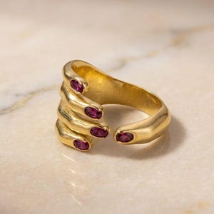 May include: A gold-toned open ring shaped like a hand with fingers wrapping around the band. Each fingertip is accented with a small, oval-shaped, deep pink gemstone. The metal has a smooth, polished finish and the design features five distinct finger-like extensions.