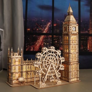 May include: A wooden model of London landmarks, including Big Ben, the London Eye, and a building with a bench. The model is made of light-colored wood and has intricate details. The structures are illuminated from within, creating a warm glow.