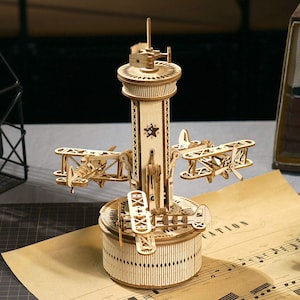 Op de afbeelding: Een houten model van een carrousel met dubbeldekkers. Het model heeft een centrale toren met een sterembleem, een draaiende basis en verschillende kleine dubbeldekkers die aan armen zijn bevestigd. Het model is gemaakt van lichtgekleurd hout.