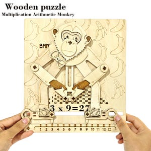 May include: Wooden multiplication arithmetic monkey puzzle. The light-coloured wooden puzzle features a monkey design with movable arms and legs. The puzzle includes multiplication problems and banana illustrations. The text "3 x 9 = 27" is visible.