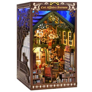May include: A miniature wooden diorama of a cozy cat-themed room, illuminated with warm lighting. The diorama features a detailed interior with bookshelves, furniture, and the text "Cat Affairs Bureau" at the top. The exterior is a dark brown with a tree silhouette.