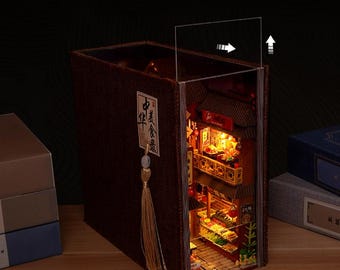 Op de afbeelding: Een miniatuur, verlicht Chinees restaurant diorama in een boekvormige behuizing. Het gedetailleerde model heeft een rood dak, balkons, voedseldisplays en warme binnenverlichting. Een bruine kwast hangt aan de zijkant.