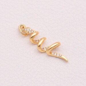 Op de afbeelding: Een goudkleurige metalen hanger in de vorm van een opgerolde slang. Het lichaam van de slang is voorzien van een reeks kleine, heldere, fonkelende steentjes en aan de bovenkant zit een klein rond oogje om aan een ketting te bevestigen.
