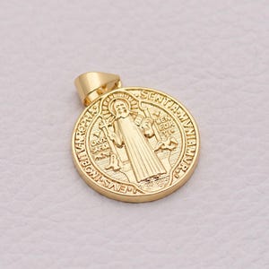 Puede incluir: Un colgante dorado con un diseño religioso detallado. El colgante redondo presenta la imagen de un santo y texto. El colgante tiene un lazo en la parte superior para una cadena. El fondo es de color claro y neutro.