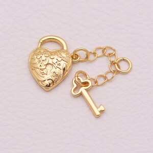 May include: A gold-toned heart-shaped padlock charm featuring an intricate floral engraving on its surface. The padlock is connected by a short gold-toned chain to a small, matching key charm with a heart-shaped bow at the top of the handle.