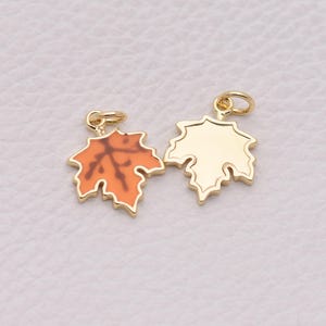 Può includere: Due ciondoli in metallo color oro a forma di foglie d'acero. Uno presenta un inserto in smalto arancione liscio con un motivo a venature marrone scuro, mentre l'altro è in metallo color oro semplice con un bordo in rilievo e un piccolo anellino.