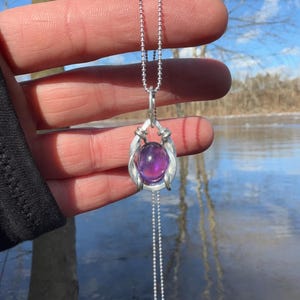 May include: A silver chain necklace with a purple amethyst gemstone pendant. The pendant is set in a silver wire cage.