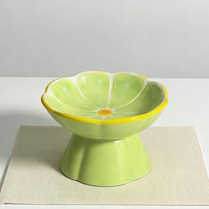 Peut inclure: Un bol en c&eacute;ramique vert citron en forme de tranche d'agrume, dot&eacute; d'un bord festonn&eacute; avec une bordure jaune, un cercle orange central et des lignes radiales blanches. Le bol repose sur une base pi&eacute;douche large et &eacute;vas&eacute;e au fini brillant.